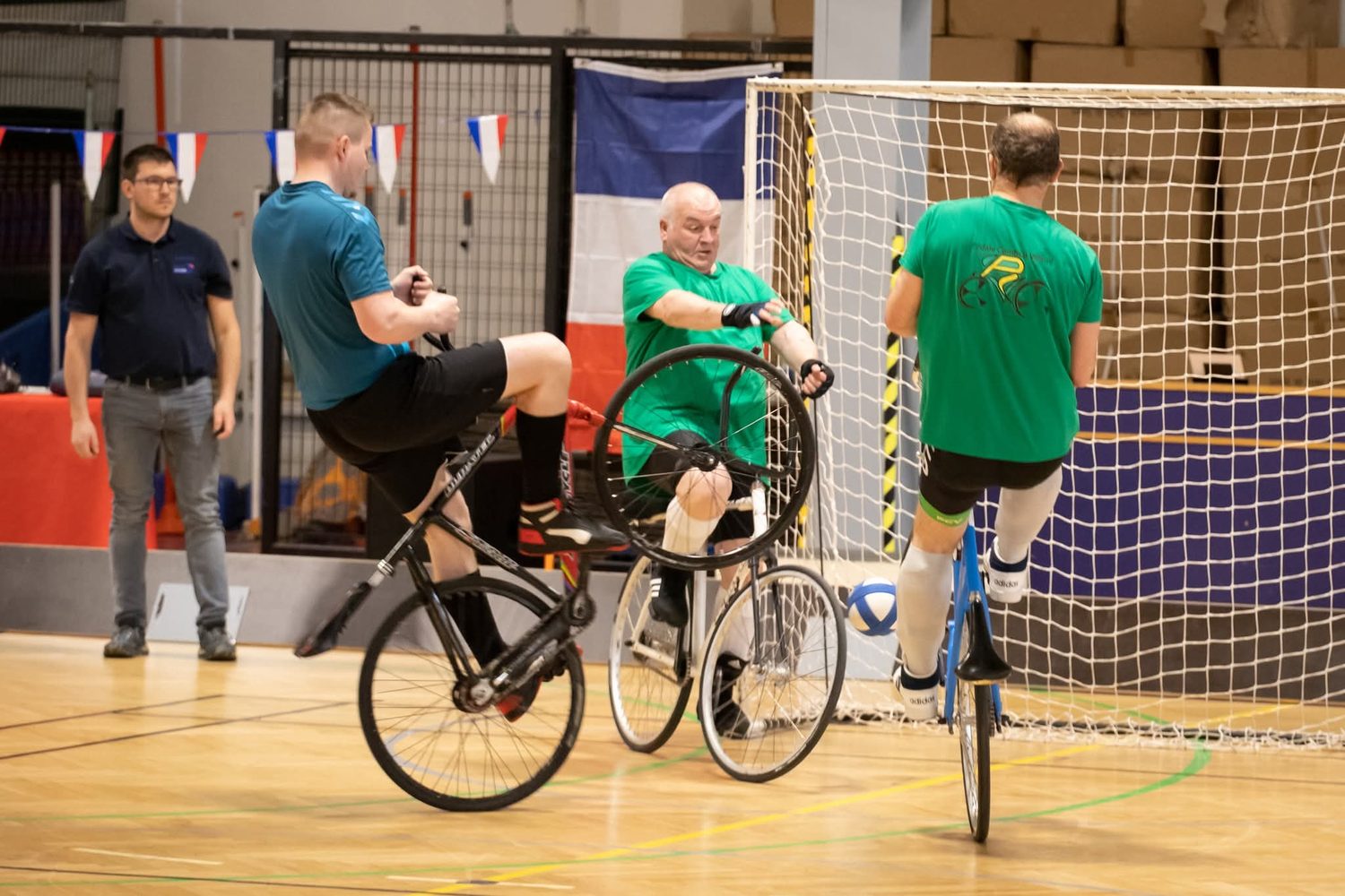 Match de cycle-balle : deux joueurs en vert défendent leur but pendant qu'un joueur en bleu attaque, tous en équilibre sur leur vélo.