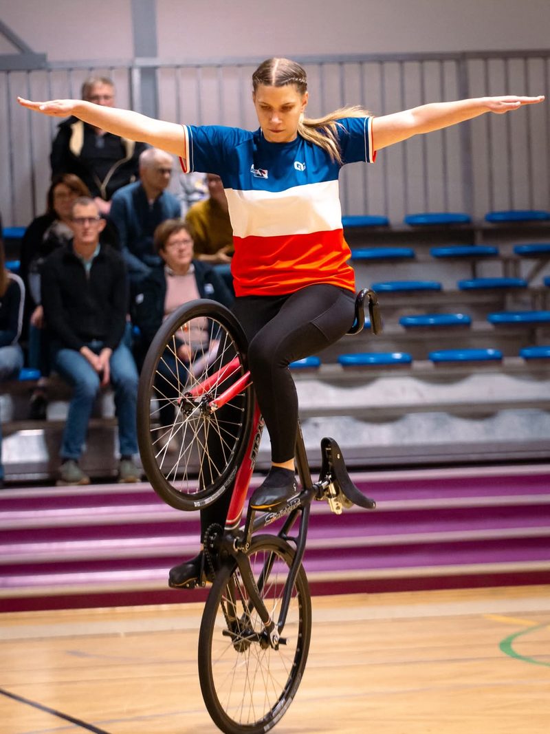Une cycliste en maillot tricolore tient une figure d'équilibre sur son vélo devant les gradins d'un gymnase.